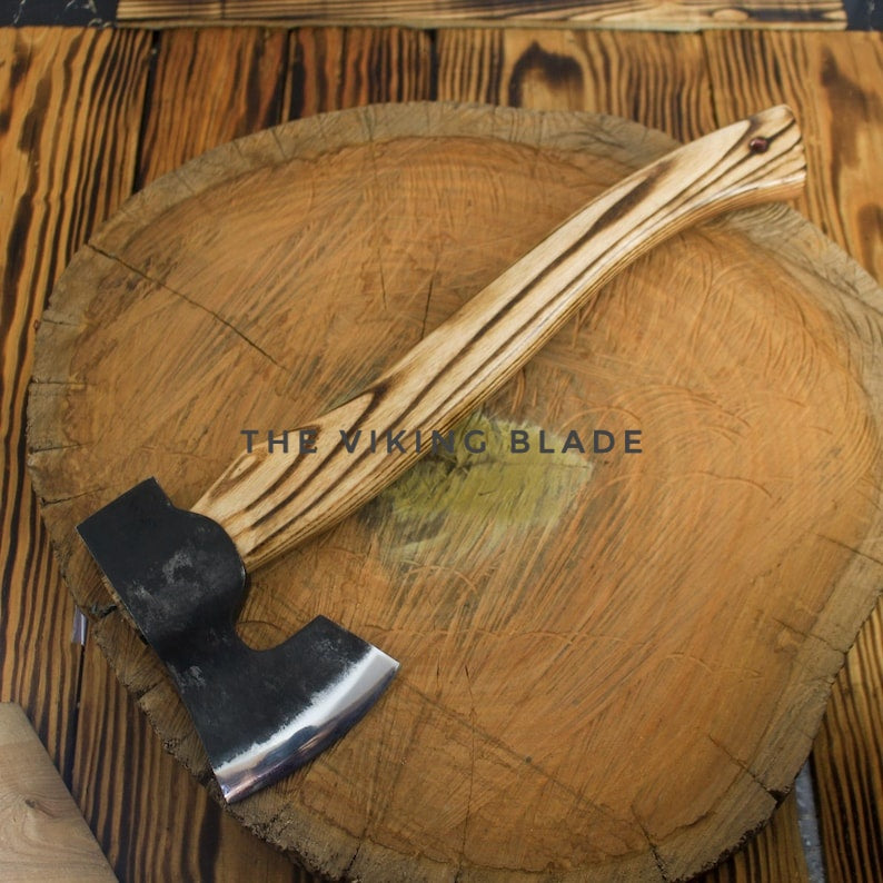 A hand axe with a striped light brown and dark brown wooden handle and a black steel head, resting on a large round wood slice over a dark wood background. The text 'THE VIKING BLADE' is overlaid across the center.