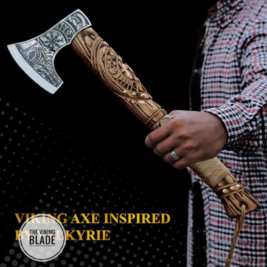 A person holding a handcrafted carbon steel Viking style axe with a decorative handle, accompanied by a leather sheath. The axe has a sharp blade and a sturdy wooden handle with a patterned design.