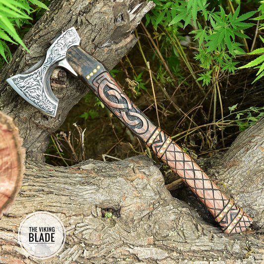 A handmade carbon steel Viking axe with a leather sheath, the axe has a decorative pattern on the handle and a Viking-themed design on the blade, placed against a natural outdoor background.