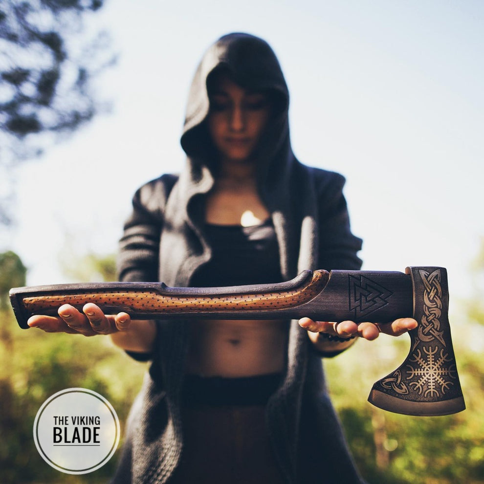 A person holding a Viking axe with a leather sheath, the axe features an engraved pattern on the blade and a plain wood handle.