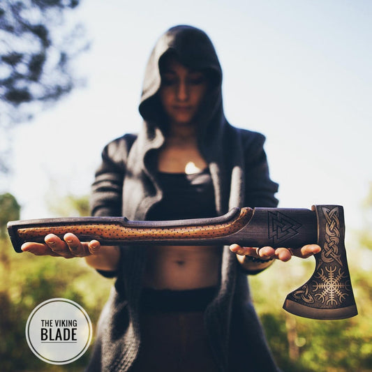 A person holding a Viking axe with a leather sheath, the axe features an engraved pattern on the blade and a plain wood handle.