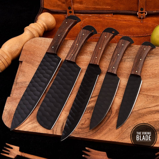 A set of custom handmade chef knives with black blades and brown wooden handles, displayed on a wooden surface with a leather roll kit in the background.