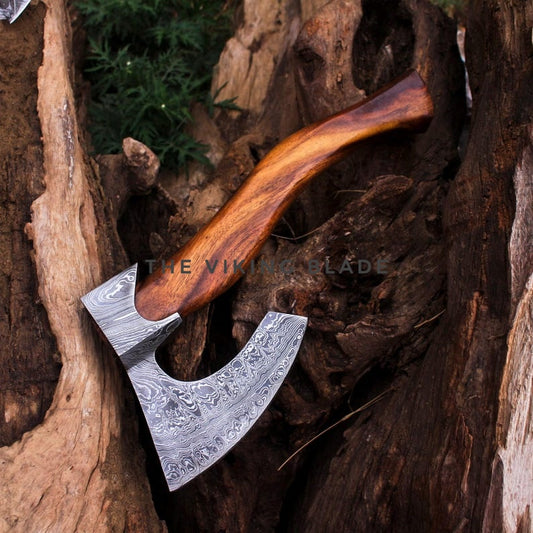 A hand axe with a patterned Damascus steel blade and a curved brown wooden handle, resting inside a hollow of weathered tree bark.