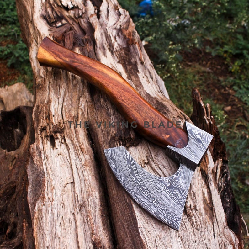 A double-bitted axe with a Damascus steel blade and a polished wooden handle, resting on a weathered log outdoors. A watermark reading 'THE VIKING BLADE' is visible across the center.