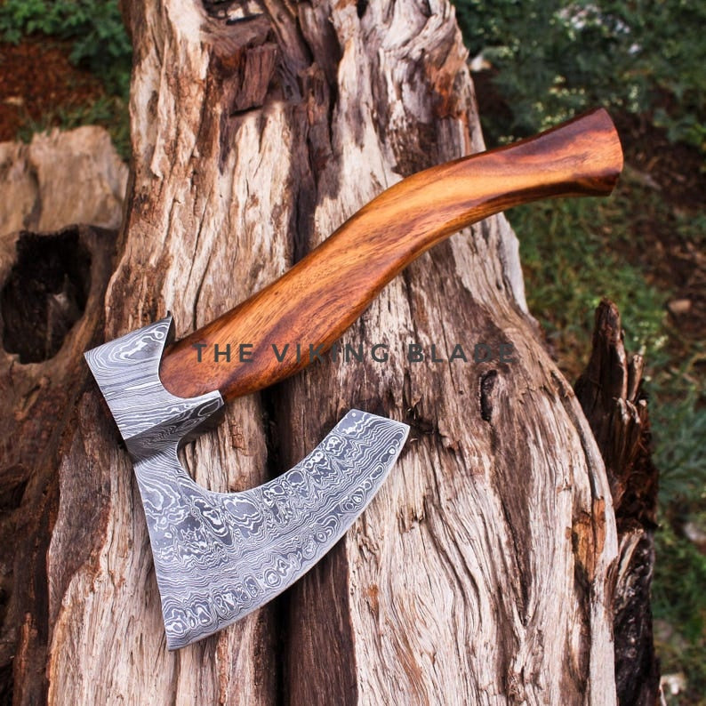 A hand axe with a patterned steel blade and a polished wooden handle, resting on a weathered tree trunk outdoors. A watermark reading 'THE VIKING BLADE' is visible across the center.