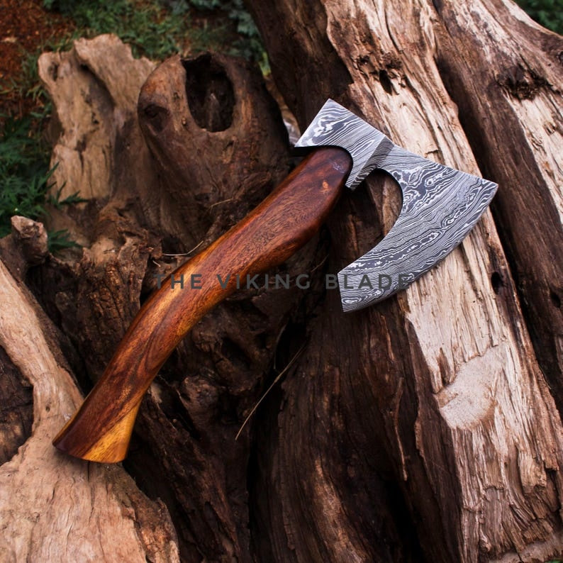 A hand-crafted Viking-style axe with a patterned Damascus steel blade and a polished brown wooden handle, resting on a textured background of natural tree bark and logs.