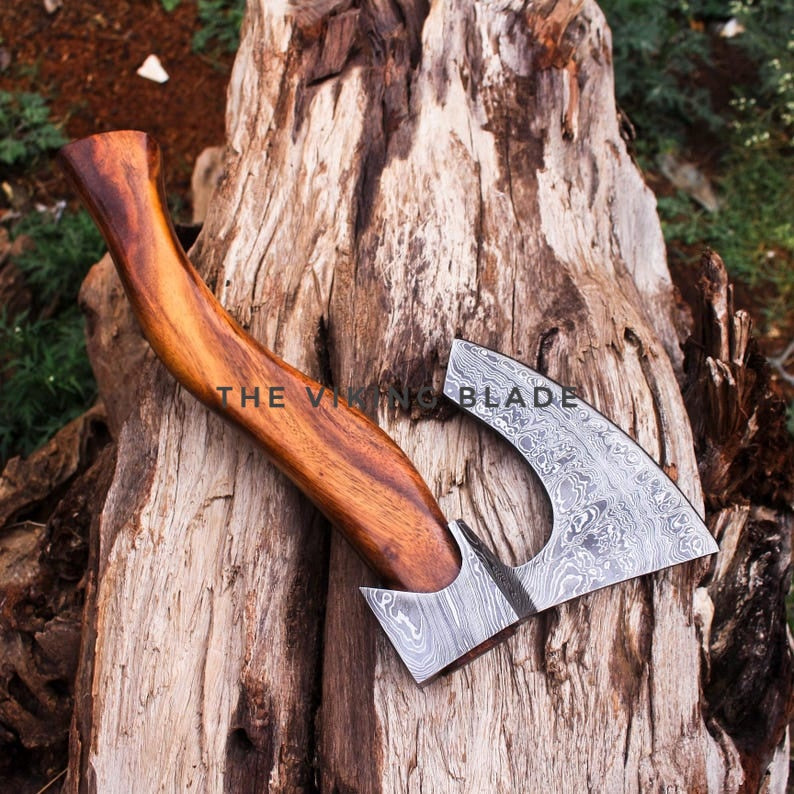 A hand-crafted Viking-style axe with a patterned Damascus steel blade and a polished wooden handle, resting on a textured log outdoors. A watermark reading 'THE VIKING BLADE' is visible across the center.