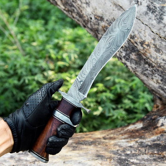 A hand holding a custom handmade Damascus steel Bowie knife with a wooden handle and a patterned blade, in front of a natural outdoor background.