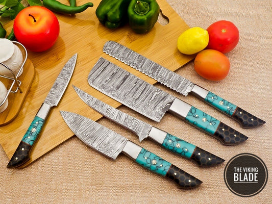 A set of custom handmade Damascus steel chef kitchen knives with colorful resin handles and a patterned steel finish, displayed alongside a leather roll kit, on a wooden surface with vegetables in the background.