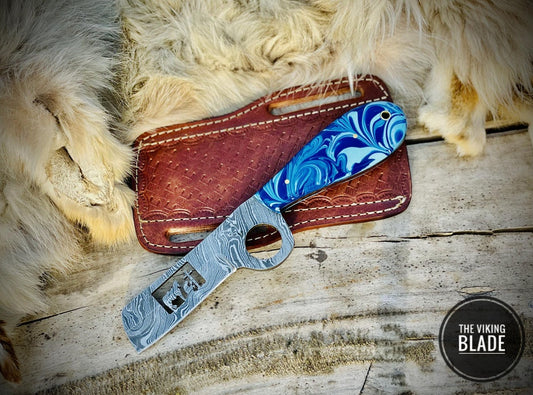 A custom handmade Damascus steel cowboy bull cutter knife with a blue-colored resin handle, displayed alongside its brown leather sheath on a wooden surface.