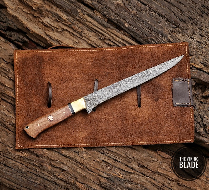 A custom handmade Damascus steel fillet fish chef knife with a wooden handle featuring a brass guard and shark mosaic, resting on a brown leather roll kit.