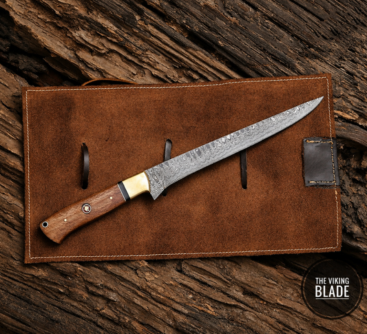 A fixed-blade knife with a patterned steel blade, a brass bolster, and a brown wooden handle, displayed diagonally on a brown leather sheath over a rustic wood background. A circular logo in the bottom right corner reads 'THE VIKING BLADE'.