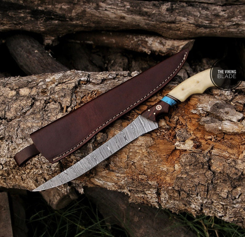 A custom hand forged Damascus steel fillet knife with a brown leather sheath, resting on a wooden surface with a natural outdoor background.