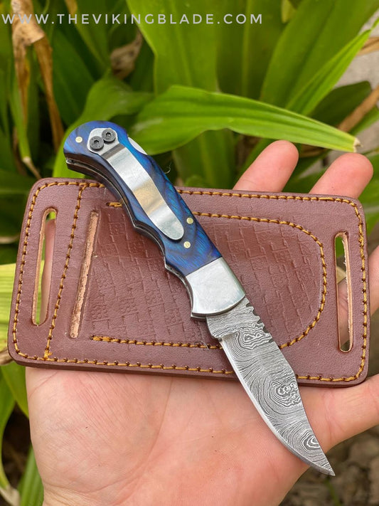 A handmade Damascus steel folding pocket knife with a blue and silver handle, placed on a brown leather sheath, held in a person's hand.
