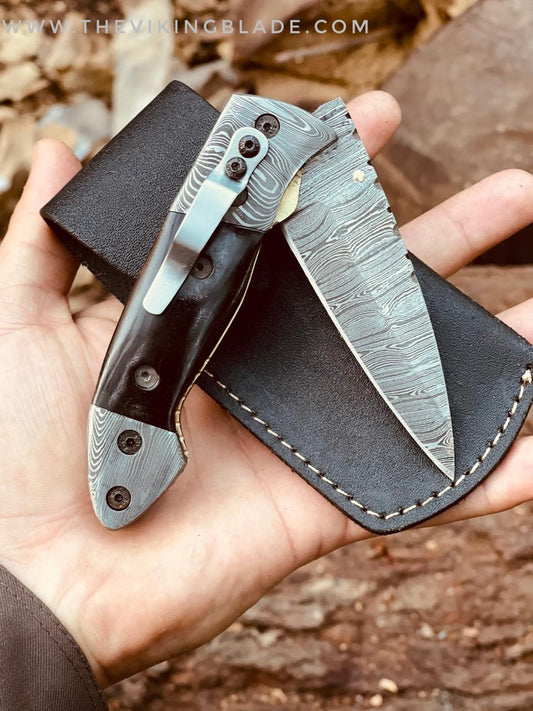 A handmade Damascus steel folding pocket knife with a leather pouch. The knife is open with the blade facing up, and the pouch is closed with the knife partially inserted.
