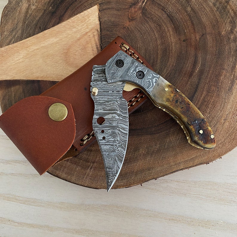 A Damascus steel folding pocket knife with a brown handle, partially opened, resting on a wooden surface with a leather sheath beside it.