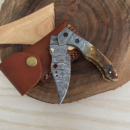A Damascus steel folding pocket knife with a brown handle, partially opened, resting on a wooden surface with a leather sheath beside it.