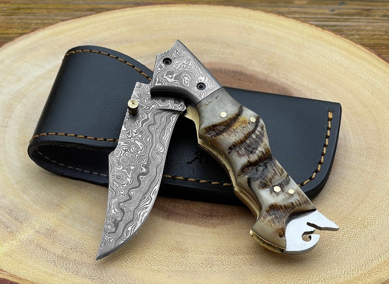 A Damascus steel folding pocket knife with a ram horn handle, displayed alongside its leather pouch sheath.