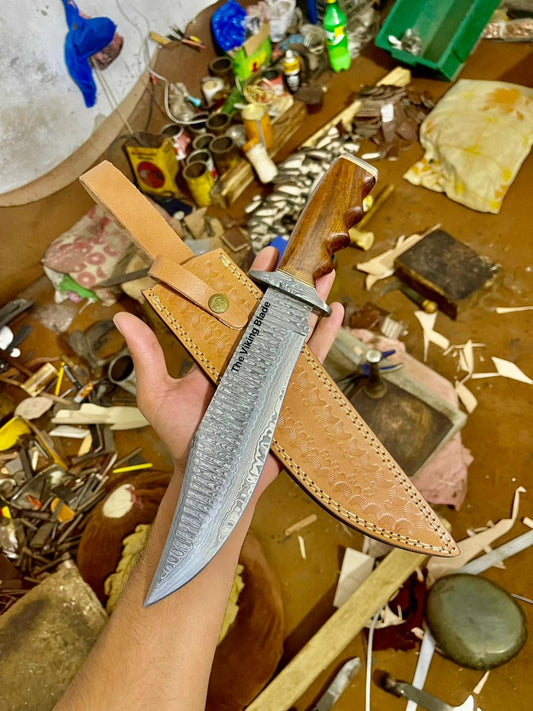 Handmade Damascus steel hunting Bowie knife with a leather sheath, held in a person's hand, with various other objects in the background.