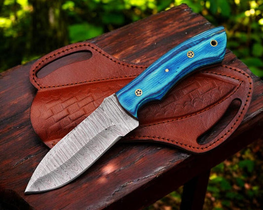 A custom handmade Damascus steel hunting knife with a blue and red leather sheath, placed on a wooden surface in a natural outdoor setting.