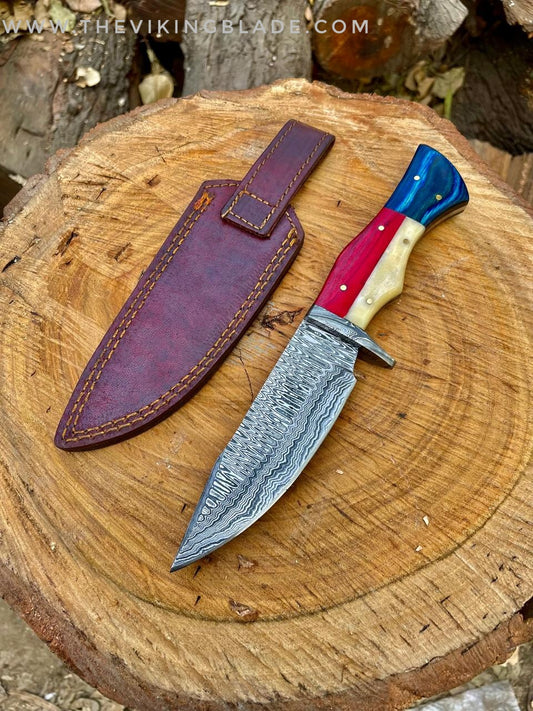 A handmade Damascus steel hunting knife with a brown leather sheath, displayed on a wooden surface.