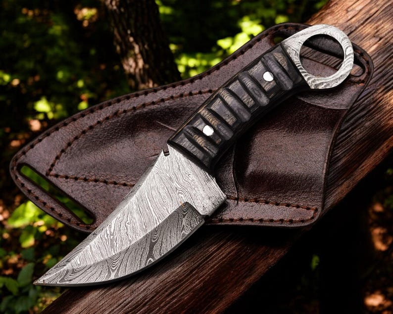 A Damascus steel skinner knife with a black and gray Pakka wood handle and a leather sheath, placed on a wooden surface in a natural outdoor setting.