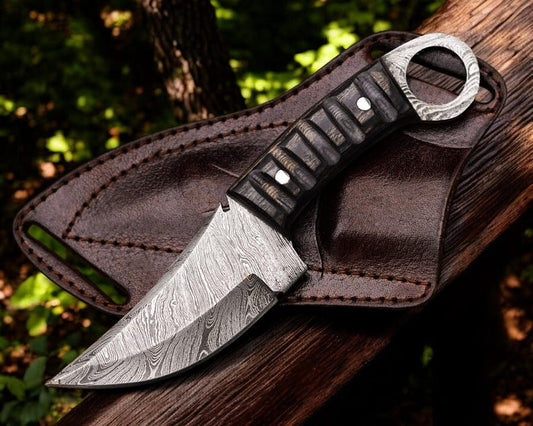 A Damascus steel skinner knife with a black and gray Pakka wood handle and a leather sheath, placed on a wooden surface in a natural outdoor setting.