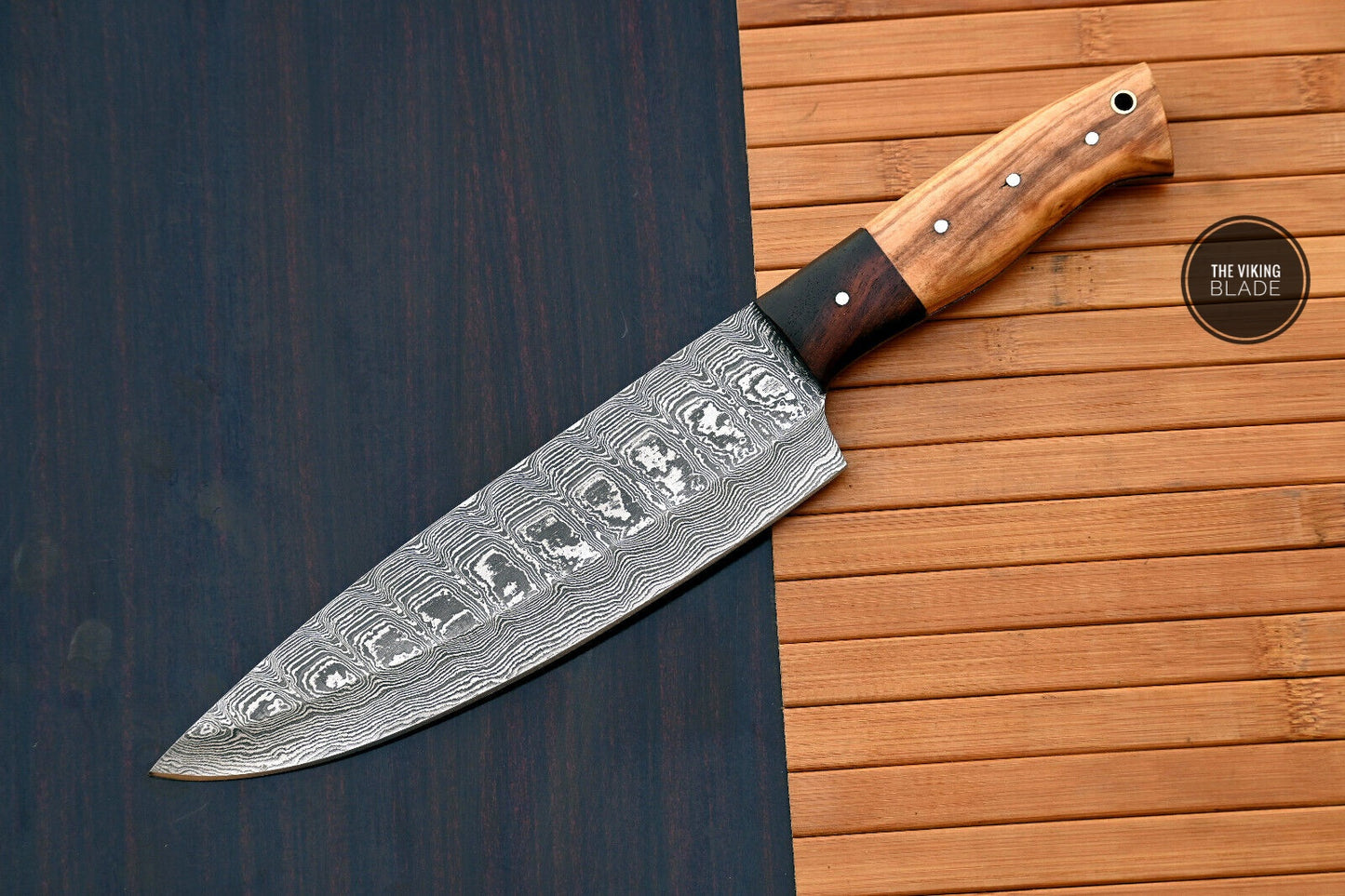 A hand forged Damascus steel chef knife with a wooden handle and a patterned blade, resting on a wooden surface.