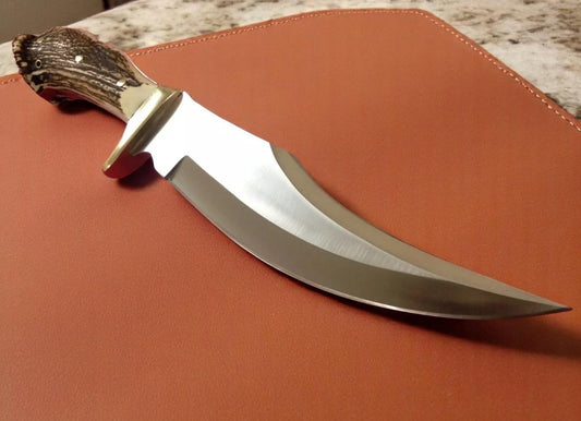 A custom handmade hunting Bowie knife with a blade material of D2 Tool Steel and a handle material of Crown Stag, featuring a brass finger guard. The knife is displayed on a brown leather sheath.