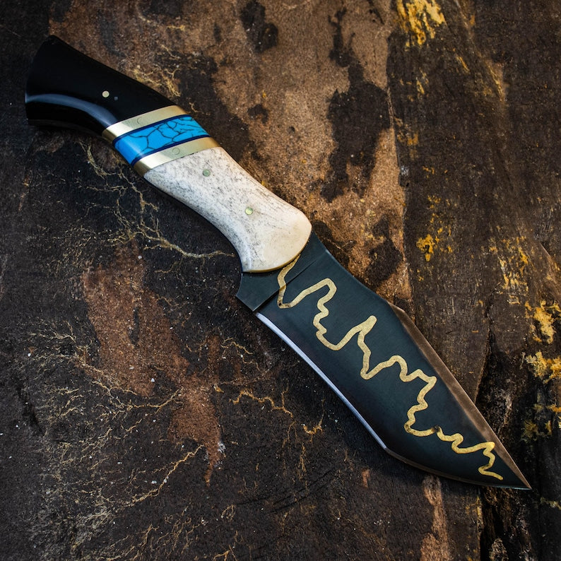A custom handmade hunting Viking knife with a black blade featuring a golden pattern, and a handle with a black and white grip, placed on a textured dark background.
