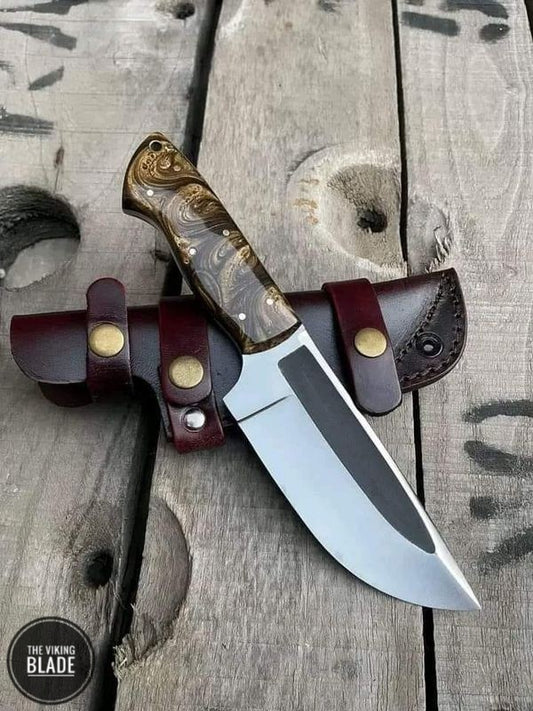 A custom handmade Skinner hunting knife with a D2 steel blade and a brown leather sheath, displayed on a wooden surface.