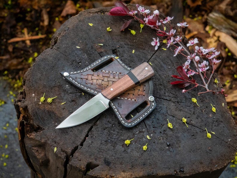 A custom handmade hunting Bowie knife with a sheath, placed on a wooden stump surrounded by leaves and flowers.