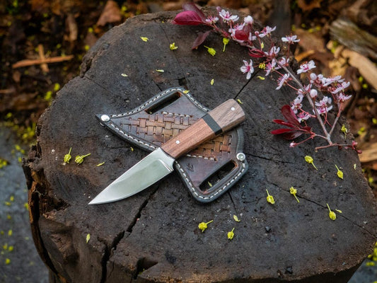 A custom handmade hunting Bowie knife with a sheath, placed on a wooden stump surrounded by leaves and flowers.