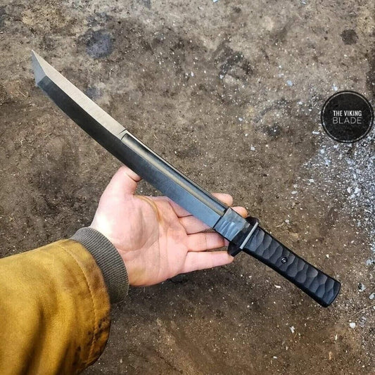 A hand holding a custom handmade Spring Steel 5160 Hunting Tanto Knife with a black handle and a leather sheath.
