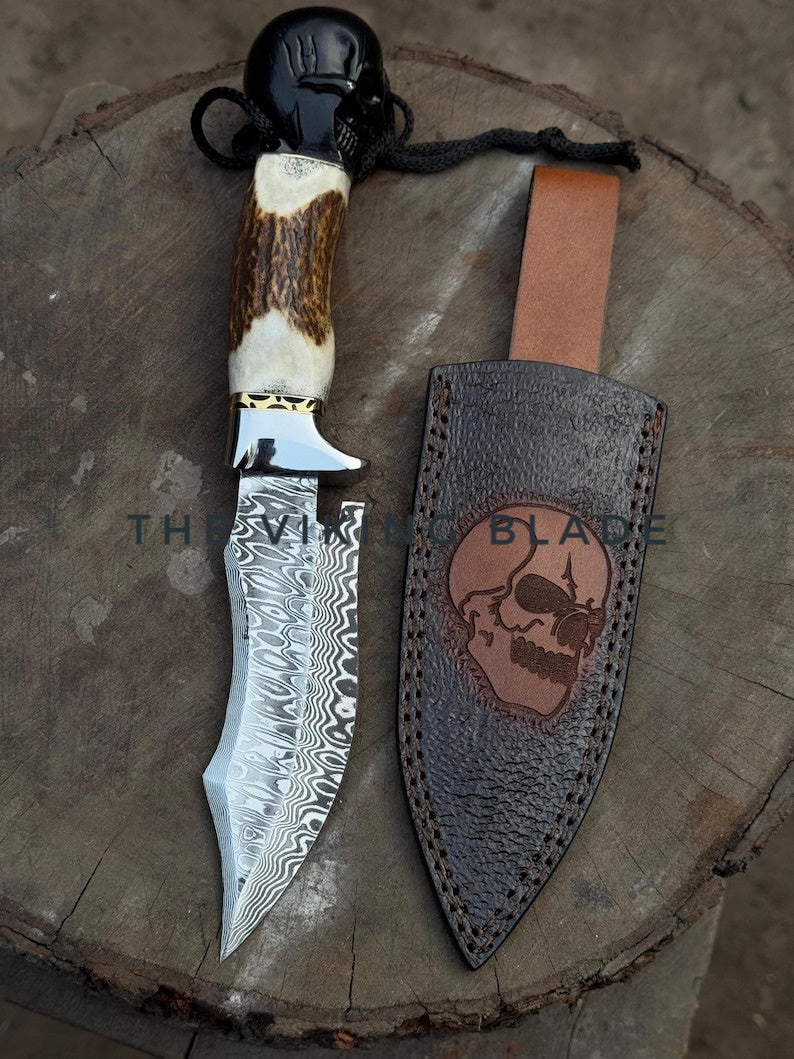 A stainless Damascus steel hunting knife with a patterned blade, deer horn handle featuring a black skull pommel, and brass guard. The knife is accompanied by a handcrafted leather sheath with skull engraving.