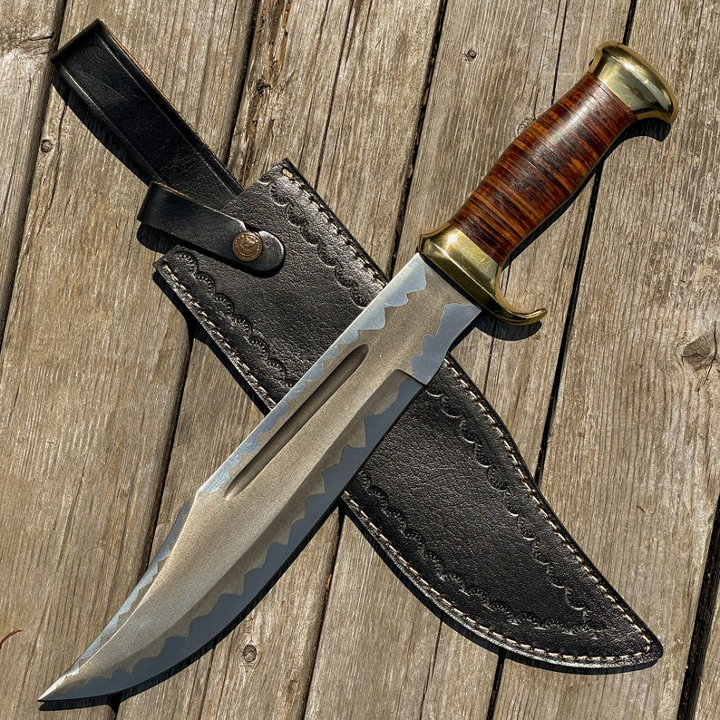 A custom handmade stainless steel hunting Bowie knife with a leather sheath, featuring a wood and leather handle with brass accents on a wooden background.
