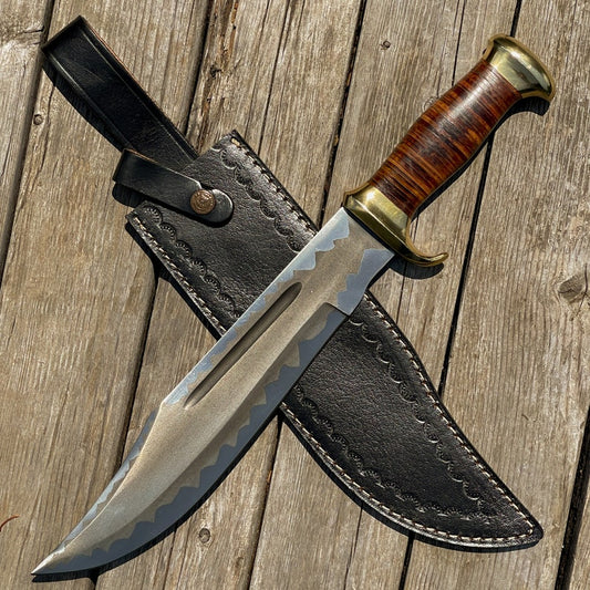 A custom handmade stainless steel hunting Bowie knife with a leather sheath, featuring a wood and leather handle with brass accents on a wooden background.
