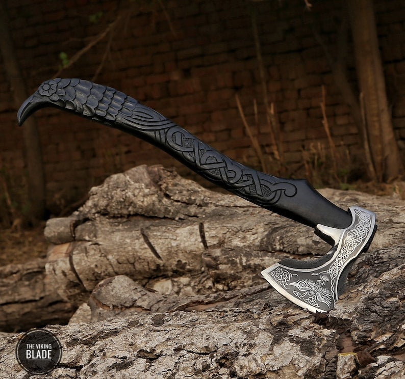 A hand-forged carbon steel Viking axe with a black leather-wrapped handle featuring Norse mythology-inspired engravings, resting on a wooden stump with a natural background.