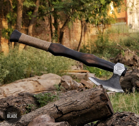 A handmade Viking axe with a carbon steel head and an ash wood/rose wood handle, displayed with a leather sheath beside a wooden stump in a natural outdoor setting.