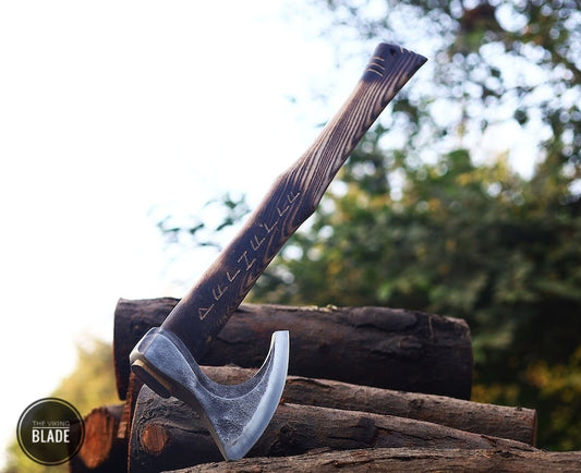 A custom handmade Viking axe with a carbon steel head, ash wood handle, and a leather sheath, placed on a wooden surface with a blurred background.
