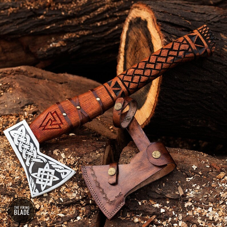 A Viking axe with a carbon steel blade and an ash wood handle featuring a patterned design, accompanied by a brown leather sheath, placed against a natural wood background.