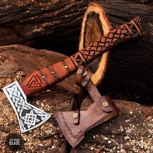 A Viking axe with a carbon steel blade and an ash wood handle featuring a patterned design, accompanied by a brown leather sheath, placed against a natural wood background.
