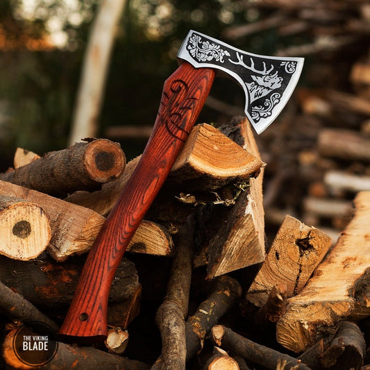 A handmade hatchet with a red wooden handle and a black leather sheath, placed on a pile of wooden logs.