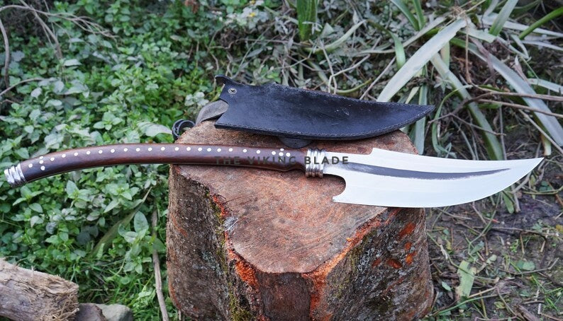 A custom handcrafted knife with a curved silver blade, a brown handle with silver studs, and a black sheath, displayed on a rustic tree stump outdoors.