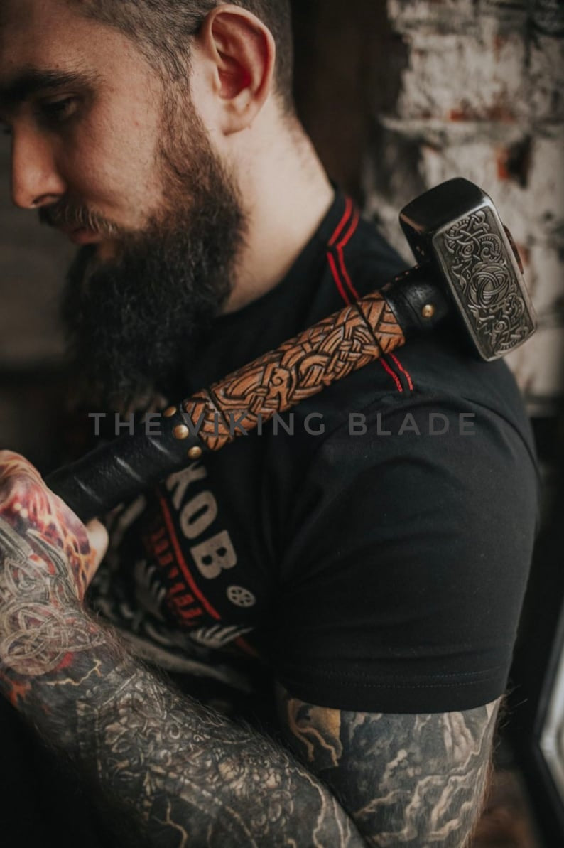 Man with tattoos wearing a black shirt with visible branding, holding a decorative hammer.