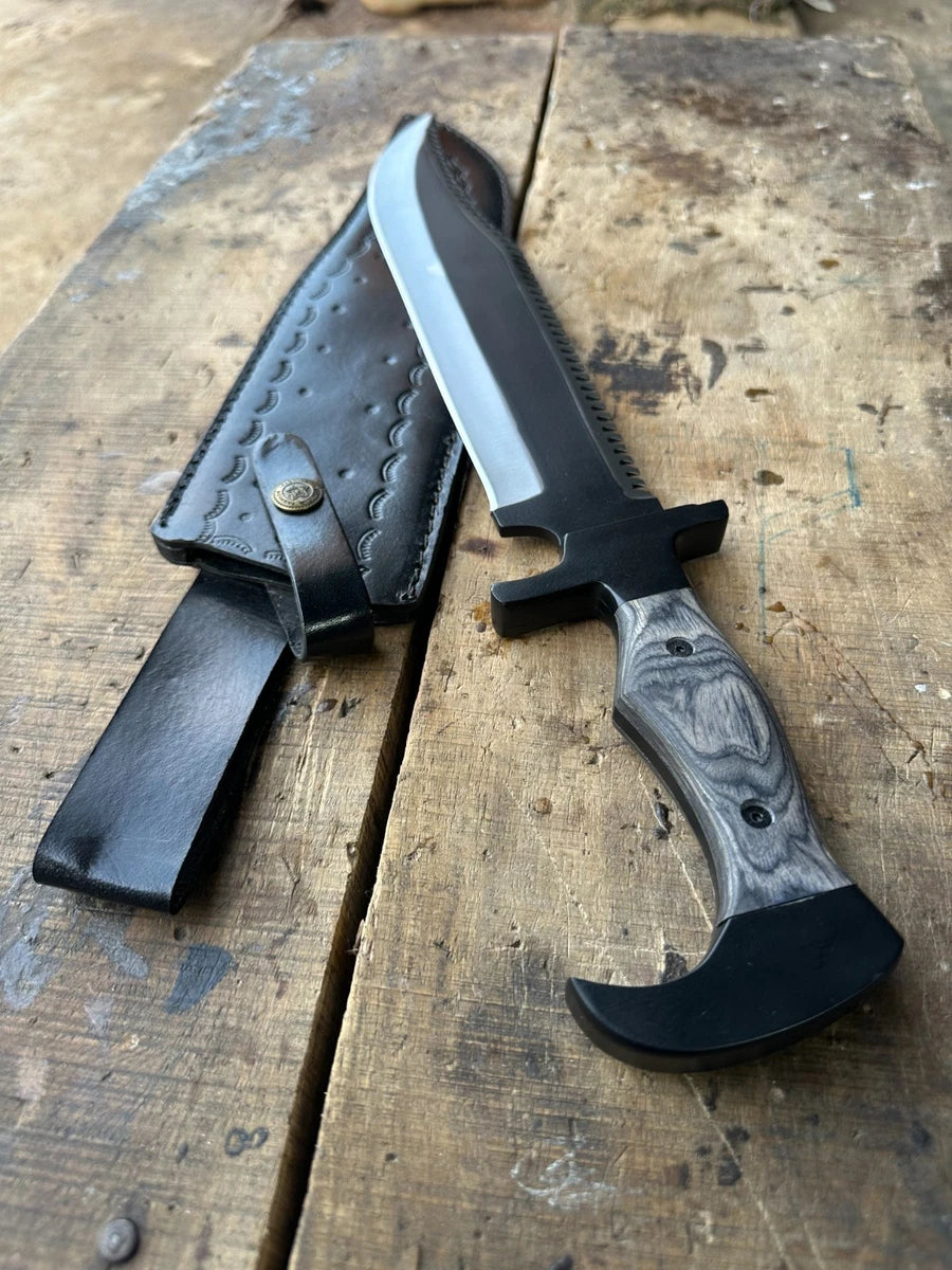 A fixed-blade knife with a black blade, black handle, and gray marbled handle scale, shown next to a black leather sheath on a rustic wooden surface.