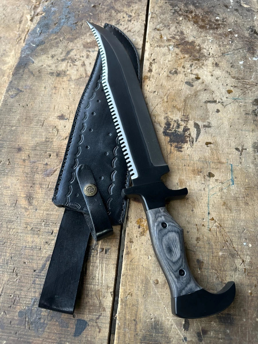 A fixed-blade knife with a black blade, serrated edge, black handle, and a gray wood-grain handle scale, shown next to its black leather sheath on a rustic wooden surface.
