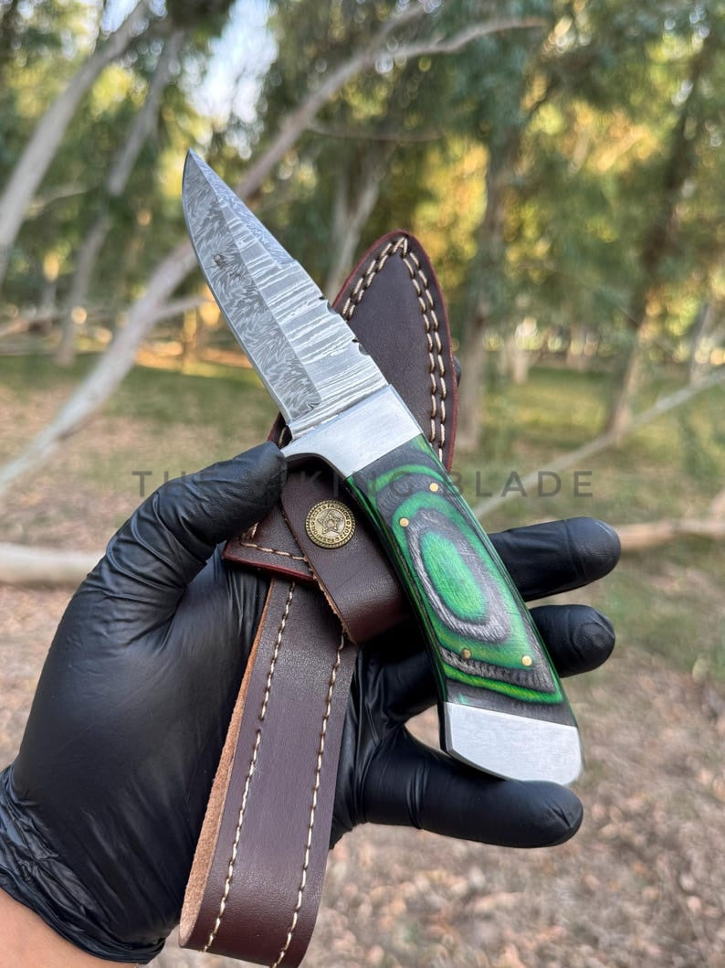 Knife with green handle and brown leather sheath held by a gloved hand against a natural background.