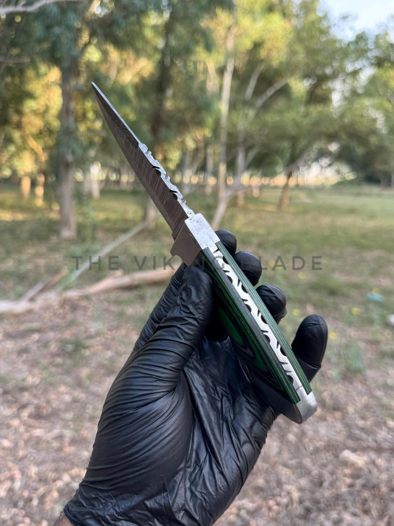 Hand holding a knife with a green blade and black handle, outdoors.