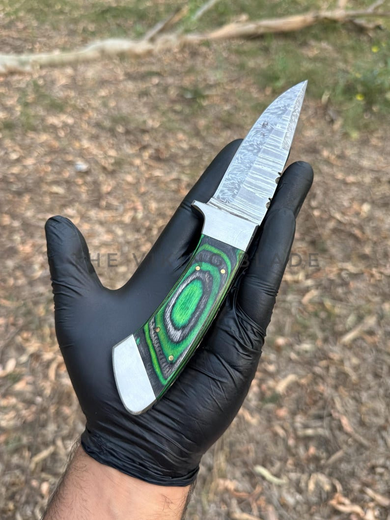 Hand wearing a black glove holding a knife with a green handle against a natural background